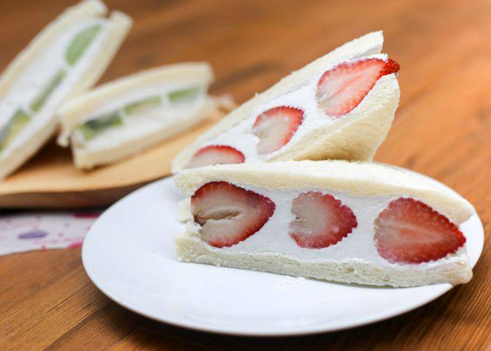 Two halves of a strawberry and whipped cream sandwich. In the background, a kiwi and cream sandwich.